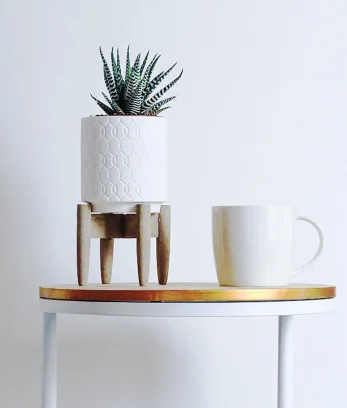 A white table with a gold top against a white wall. There is a potted plant and mug on top of the table.
